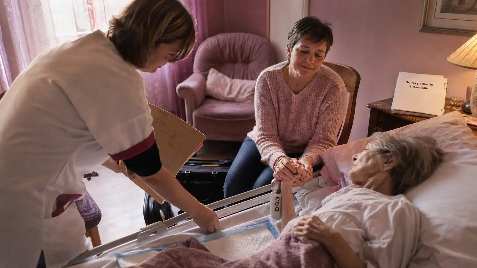 Soin palliatif domicile : infirmière ajuste un lit médical, proche tient la main du patient